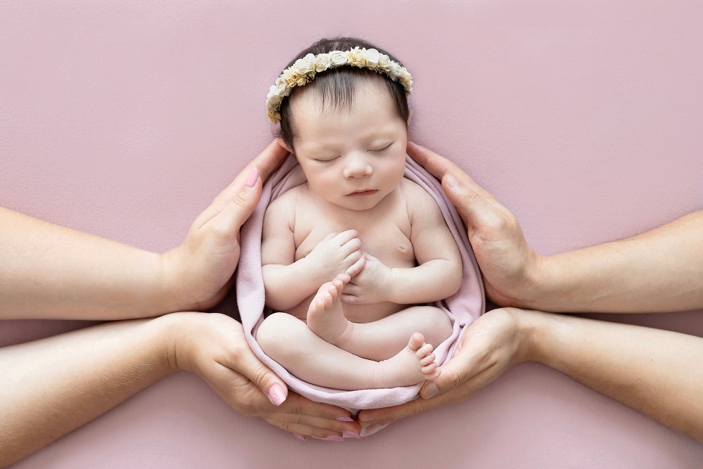 Newborn baby girl in parents’ hands wearing floral headband on pink blanket, Essex photographer